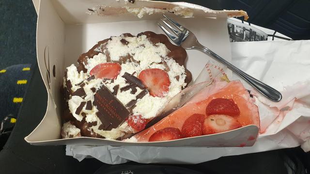 Auf meinem Schoß liegt eine kleine Pappschachtel, die eine Waffel mit einem Berg Sahne obendrauf und ein Stück Erdbeertorte mit einer Basis aus Quark o.ä. enthält. Beides ist schon leicht angedrückt, sieht aber immer noch recht nobel aus.
Auf dem Sahneberg der Waffel liegt zwischen einigen Erdbeerscheiben ein bedrucktes Schokoladentäfelchen, welches verrät, dass der Inhalt der Schachtel von Rene's Croissants Amsterdam stammt.
Mein Gäbelchen liegt bereit daneben, es kann losgehen.
