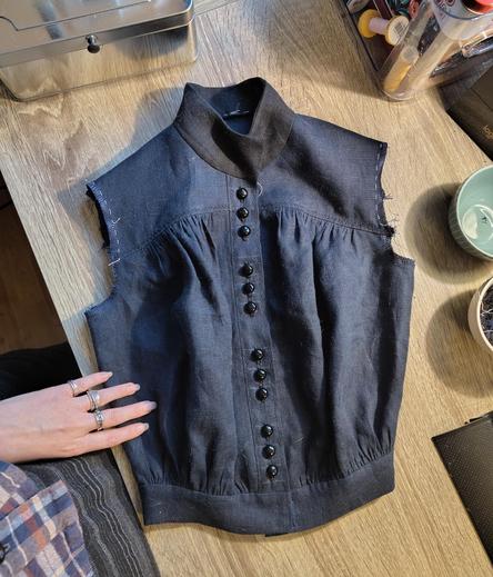 A dark blue linen shirtwaist on a desk, sans sleeves, but otherwise looking fairly finished. There's twelve small shiny black buttons of a dome shape on the button placket, in groups of three, very closely spaced. There are no buttons on the black linen collar or the blue waistband. The collar flops about freely while the waistband is held closed. There's a lot of blousing from gathered fabric at the bust and waist, in a vaguely Edwardian shape, and the fit of the shirt is very old fashioned with sloping, narrow shoulders.